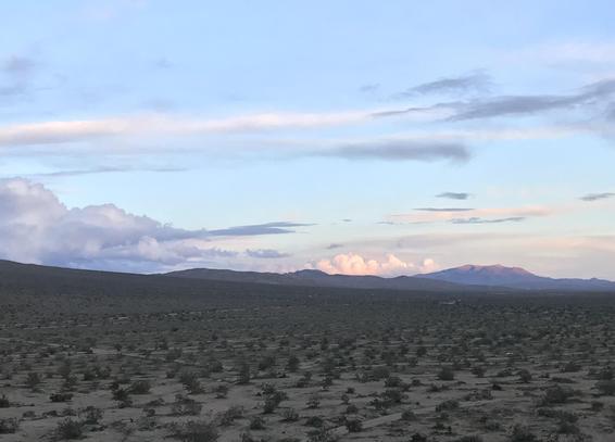 Lower half of the photo shows a sand-and-sagebrush desert, taken from a vantage point 30 feet or so above the ground. It's past sunset but not yet dark. There are low hills in the distance, the tops of which are catching the last sunlight. The top half of the frame is pale blue sky and clouds varying from gray to peach colored.