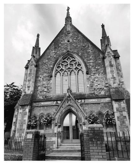 Black and white portrait photograph showing the front of an English methodist church built in the gothic style. On top of a spire at the front sits a seagull.