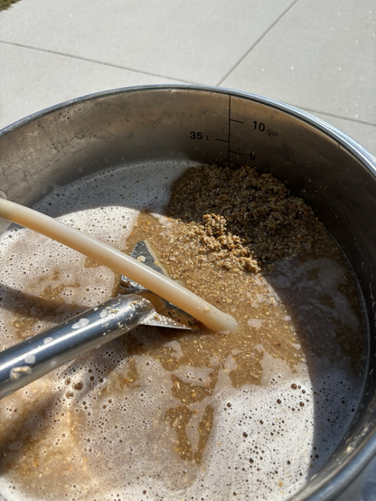 A large steel kettle is full of a hot, soupy mix of water and grain.  A hose is going into the pot, delivering more hot, delicious wort.  A paddle is also stuck into the pot, lifting some delicious grain mash out of the wort