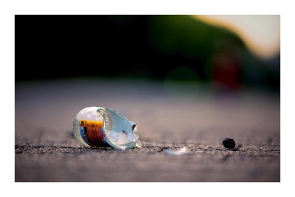 Broken glass bottle lying on asphalt with small shards scattered nearby. The background is blurred with greenery and a soft gradient of light.