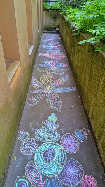 Das Bild zeigt eine schmale, asphaltierte Gasse, die von zwei Wänden eingerahmt wird. Die Wände sind aus Beton und haben eine braune Farbe. Auf dem Asphalt sind farbenfrohe, handgezeichnete Muster und Designs in verschiedenen Farben wie Blau, Pink, Gelb und Grün zu sehen. Diese Muster bestehen aus Blumen, Blättern und geometrischen Formen, die in einer kunstvollen und farbenfrohen Weise angeordnet sind. Die Gasse führt zu einer Treppe, die in den Hintergrund führt, wo grüne Pflanzen sichtbar sind. Die Wand rechts ist mit Moos bedeckt, was auf eine feuchte Umgebung hinweist. Die gesamte Szene ist von einer natürlichen und künstlerischen Atmosphäre geprägt.