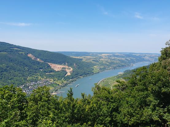 Foto vom sonnen beschienenen Mittelrheintal bei hellblauen Himmel mit Blick über den grünen Wald auf den hellblauen Rhein mit 2 Schiffen, der nach rechts fließt. Links sind ein paar Gebäude von Trechtingshausen erkennbar und großflächig bewaldete Hänge und ein großer Steinbruch