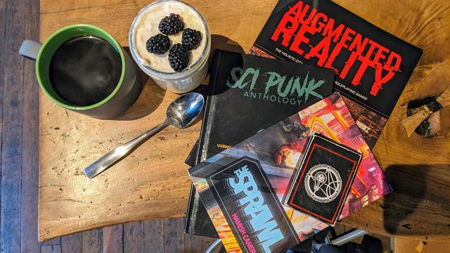 A table with a stack of rpg books - The Sprawl, Sci Punk, and Augmented Reality - with a cup of black coffee, and overnight oatmeal with blackberries beside them.