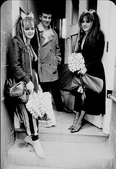 At the left Jill stands on the top step in front of the open door, bow in her hair, thick eyeliner, polka dot umbrella and briad vertically striped trousers. At the right, Rose leans against the open door wearing a long black coat, also with a bow in her hair and polka dot umbrella. Between them, and slightly further away across the threshold of the doorway, stands Drew in a sheepskin coat. They are all looking at the camera, with the impression that this was a spontanteous shot.