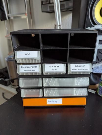 Ein Foto eines kleinen, selbst gedruckten Sortimentskasten. Er ist schwarz und hat Fächer für Schubladen in unterschiedlicher Größe. In einigen Fächern befinden sich bereits Schubladen aus transparentem oder orangem Plastik, einige sind mit Zetteln beschriftet.