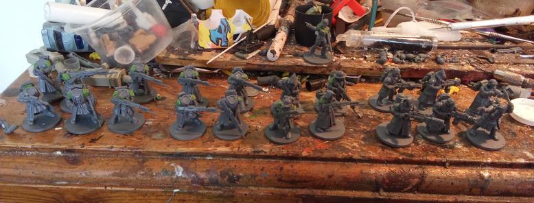 A very messy modeling table with 21 assorted models in varying states of completion. The theme is longcoats, rifles, and green stuff modifications.