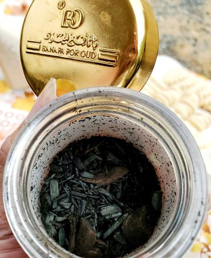 Jar of Bakhoor Oud Moattar Bahraini wood burned incense scents in small glass jar with gold coloured lid. Held in my hand with lid off & view of jar contents.