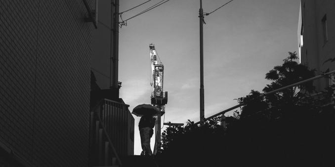 Silhouette of a person holding an umbrella, walking up a staircase between two buildings. A large construction crane is visible in the background against a dusky sky. The left side features a brick wall, and dark foliage is on the right side, adding depth to the scene.