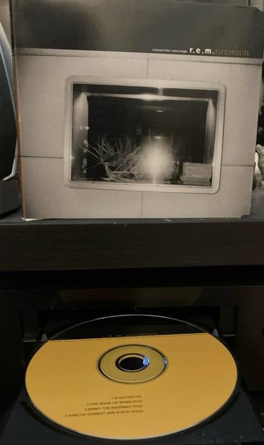 A CD case on a shelf above a CD in a player tray. The case features a black and white photograph a window at night with items on the sill. The bright spot from the flash obscures part of the window. In small type above the roof reads "compact disc maxi-single R.E.M. Electrolite"

The CD in the tray is mostly mustard yellow with a black chord at the top.  There is small writing on the disc below the spindle hole.