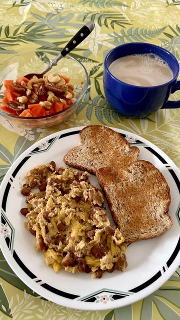 A plate with scrambled eggs, beans, and toast, next to a bowl of fruit with nuts and a huge coffee mug.