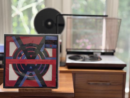 Churches - The Bones of What You Believe LP cover.

A black-bordered red square with quarter cut-outs, with a symbol inside - basically, concentric circles with two As at the bottom and top, pointing at each other. A red stripe runs through the center, with the band and title.

The black LP plays on a vintage, restored Bang & Olufsen Beogram 4000 linear-tracking turntable to the right.