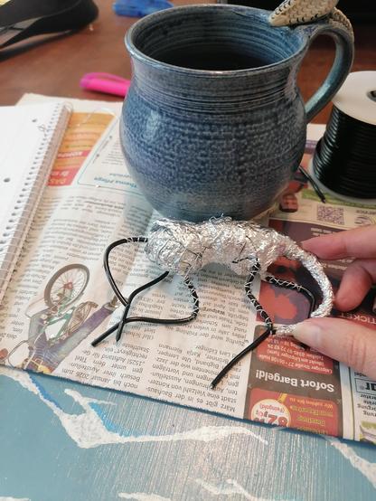 Foto vom "Skelett" der Skulptur. Mit Alufolie umwickelter Draht. Es hat vier Beine, einen Schwanz und einen Hals den es auf den Vorderbeinen ablegt. Eine bauchige Teetasse mit Drachenkopf als Henkel steht im Hintergrund.