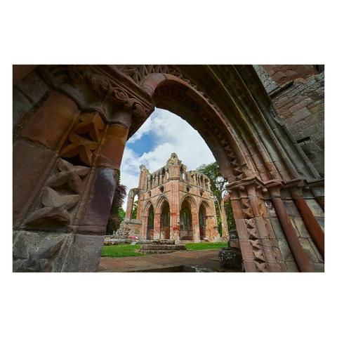 A dramatic view through a carved stone archway at Dryburgh Abbey, framing the imposing ruins of the North Transept chapel; the chapel's weathered red sandstone walls, tall pointed arches, and remnants of Gothic windows rise sharply against a bright, partly cloudy sky, with neat green grass and mature trees surrounding the ancient structure, while intricate medieval carvings are visible on the arch in the foreground.