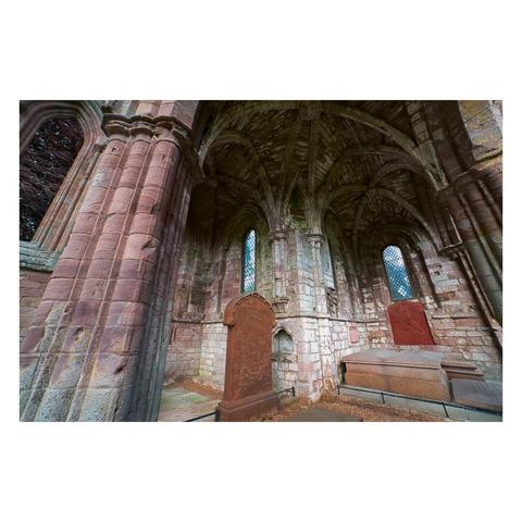 Inside the North Transept chapel of Dryburgh Abbey, the image reveals soaring ribbed stone vaults and elegant, clustered columns of weathered red sandstone, with tall lancet windows filled with diamond-patterned glass casting a gentle light across the interior; prominent gravestones and a large stone tomb rest against the walls, their inscriptions partly faded, imbuing the historic space with a reverent and timeless atmosphere.