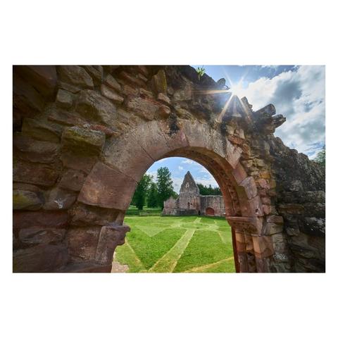 A view through a large, weathered stone archway frames the lush green grounds of Dryburgh Abbey, with sunlight streaming in from above and casting a starburst effect in the sky; directly ahead stands the refectory ruin, distinguished by its pointed gable and the remnants of a St Catherine's wheel window, surrounded by tall trees and a partly cloudy blue sky.