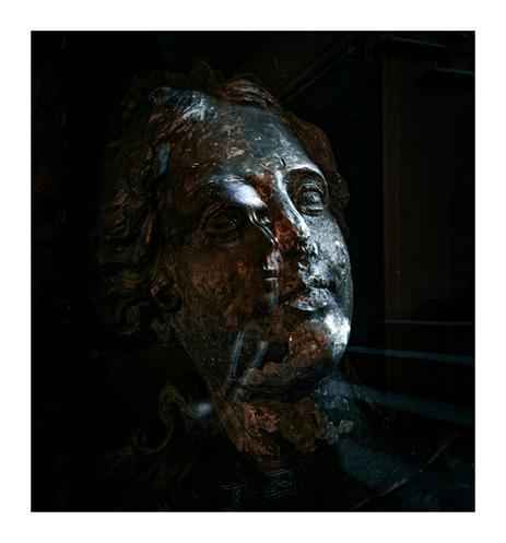 This photo shows a close-up of a weathered looking stone statue sculpture of a woman, set against a dark background.  The lighting is dramatic which highlights the texture and wear of the stone. There are strong shadows that emphasize her facial features. The bust appears aged. There is a reflection of a face superimposed on the left hand side of the statues face thanks to the glass panel. That is me, the photographer!