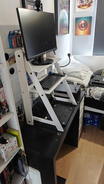 A white wooden A-frame with two shelves stands atop a black desk. It has vertical back legs and angled front legs (thus forming a right-angles triangle).
On the top shelf is a black monitor and the lower shelf holds a small keyboard. An open laptop sits on the desk surface beneath the frame.