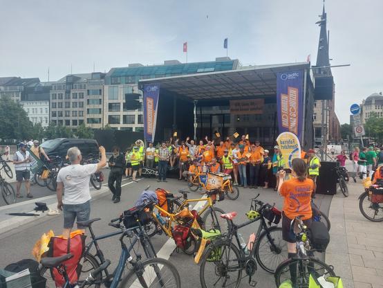 Vor einer Bühne stehen ca 20 Menschen mit Blick zum Publikum. Die meisten haben orangene T-Shirts des ADFC an. Davor steht ein orangenes Lastenrad.
