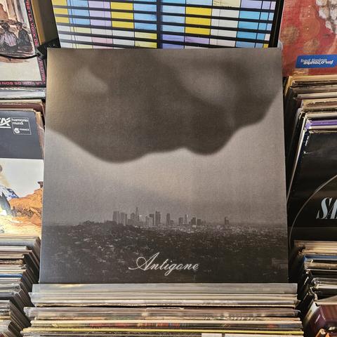 The album cover image features a black and white photograph of a  city, likely Los Angeles, in the distance, with a skyline of high-rise buildings under a cloudy, smoky sky. A dark, amorphous cloud or shadow dominates the upper portion of the cover, creating a moody and atmospheric effect. The title "Antigone" is elegantly written in a cursive font near the bottom center of the image.