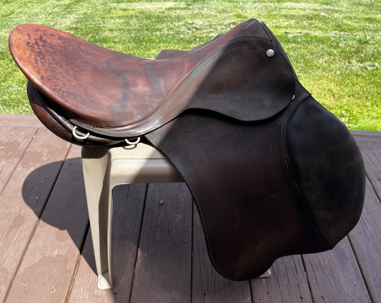 Brown English saddle drying on a tan outdoor plastic footstool. It is sitting on a brown wood deck with a grassy lawn in the background.