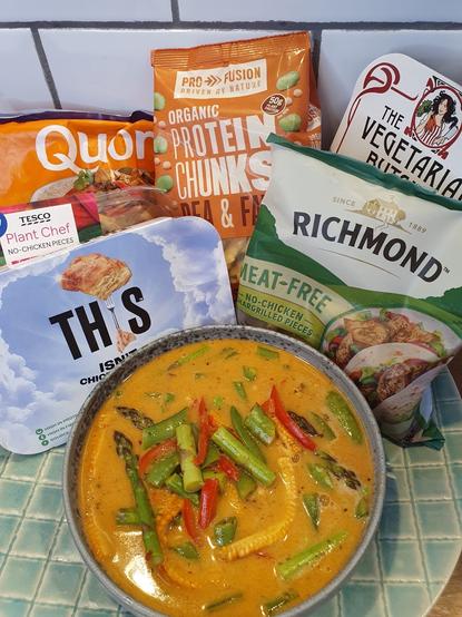 A bowl of creamy coconut curry featuring pieces of asparagus, red peppers, and baby corn, surrounded by several packages of meat alternatives, including Quorn, Pro Fusion protein chunks, and Richmond meat-free options.