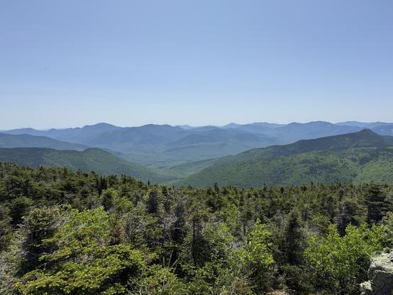 Looking down from a ridge on alpine forest as it fades downward, showing a valley with mountains on either side and a range of mountains in bluish haze along the horizon. White horizon vrsdient to light blue - perfectly clear sky. In Franconia Notch