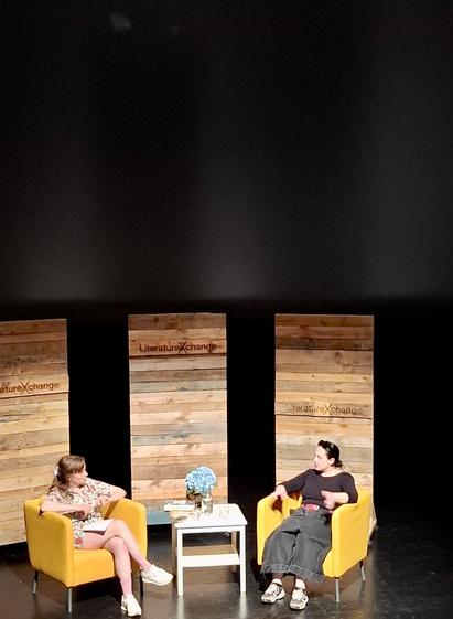 Georgian author (country not US state) Nino Haratischwili on stage in conversation with Elisabeth Skou Pedersen at LiteratureXchange Aarhus Literary Festival. They are in yellow armchairs. There are flowers, books and water on a small white side table between them. Behind them: three wooden panels with the festival name on it. Pedersen: white woman, light brown hair, wearing a beach-style summer shirt and shorts with runners. Haratischwili: white woman, black hair, wearing a smart black top and very wide-legged jeans and runners.