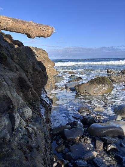 Rocky shoreline with ocean waves, scattered stones, and a large log extending over the rocks under a clear blue sky.