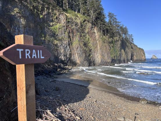 Wooden trail sign on a rocky beach with waves and tall cliffs covered in trees under a clear blue sky.