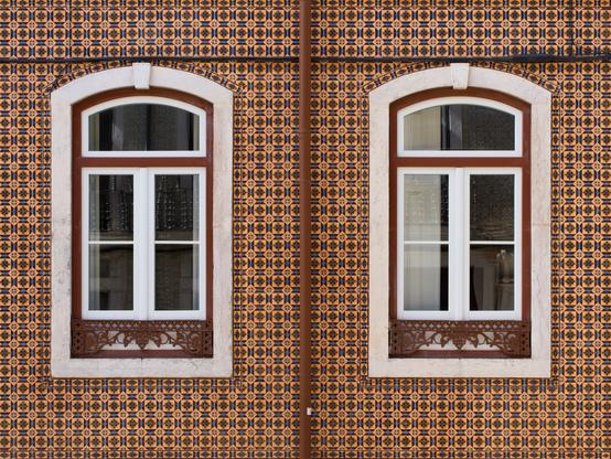 A wall of geometric ceramic tiles on a home in Lisbon, Portugal. The tiles are various shades of brown accented with bleach. A brown drainpipe runs down the center of the image and two arched windows are centered on either side of the image.