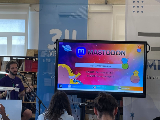 L'immagine mostra un evento in una sala con un grande schermo televisivo al centro. Sullo schermo è visualizzato il logo di Mastodon, un'applicazione di microblogging, con il testo "https://mastodon.uno" e i dati "77.000 Persone Iscritte" e "~6.000 Presenze Mensili". Sullo sfondo, ci sono due banner con il logo di Mastodon e il testo "31 VE" e "la con". A sinistra, un uomo con barba e occhiali da sole, indossa una maglietta viola e sta parlando in un microfono. Di fronte a lui, ci sono due persone con le spalle rivolte verso la telecamera, una delle quali ha i capelli lunghi e raccolti. L'atmosfera sembra informale e accogliente, con un'attenzione rivolta al presentatore e al contenuto sullo schermo.