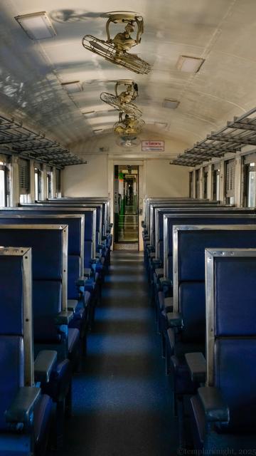 color photo in portrait format of an empty mid-century passenger train compartment with blue color upright seats on both sides and several ceiling fans on the roof, looking towards the other passenger compartments