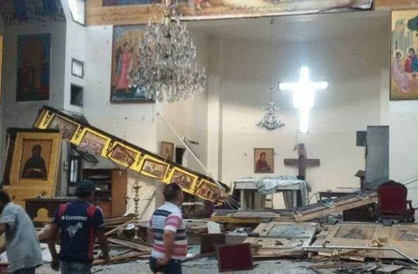 The image depicts the interior of a church that has suffered significant damage. The central focus is a large chandelier that has fallen, with its crystals scattered across the floor. The walls are adorned with religious icons and paintings, some of which are damaged or hanging askew. A prominent cross is visible on the wall, with a bright light emanating from it, suggesting it may be a window or a light fixture. The floor is littered with debris, including broken frames and pieces of wood. Several individuals are present, observing the damage. One person is wearing a striped shirt, and another has a logo on the back of their shirt. The overall scene conveys a sense of destruction and disarray within the sacred space.