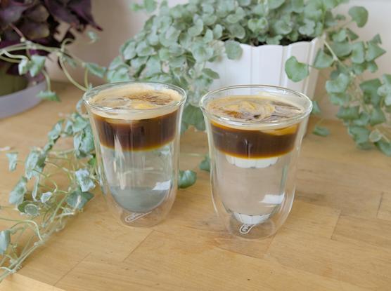 Dhá ghloine ar bhord adhmaid agus tonic espresso iontu: tá brat caife le feiscint ar barr agus líomóid is ciúbanna oighir ann, tá tonic fén gcaife. Táid potaí le plandaí ar an mbord leis.