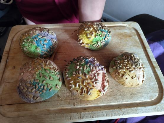 Auf dem Foto sieht man ein Holzbrett auf dem liegen fünf Brötchen die mit Lebensmittelfarbe fleckenweise in Regenbogenfarben eingefärbt wurden. Dekoriert sind sie mit Sonnenblumenkernen