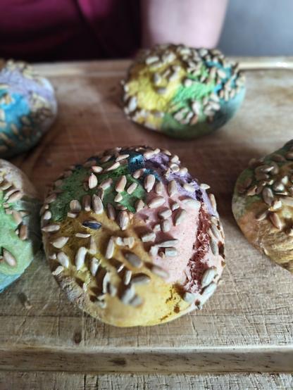 Auf dem Foto sieht man ein Holzbrett auf dem liegen Brötchen  die mit Lebensmittelfarbe fleckenweise in Regenbogenfarben eingefärbt wurden. Dekoriert sind sie mit Sonnenblumenkernen. Eins sieht man in der nahansicht.