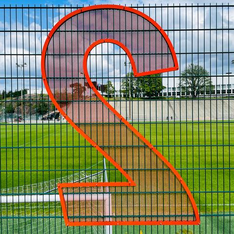 A large orange digit "2" displayed on a black mesh fence, with a green soccer field and cloudy sky in the background.