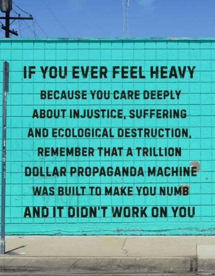 A screenshot of a post previously from Greenpeace. It’s a photo of a blue fence with black writing on top. The writing says: ‘If you ever feel heavy because you care deeply about injustice, suffering and ecological destruction. Remember that’s trillion dollar propaganda machine was built to make you numb and it didn’t work on you. ‘