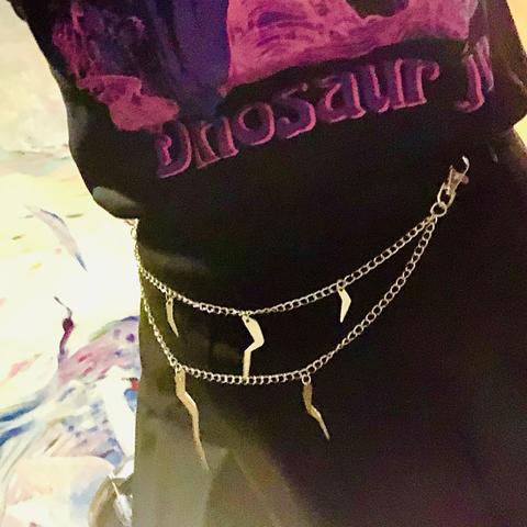 An image of a person wearing a black t-shirt featuring the text "Dinosaur Jr." adorned with a decorative chain belt that has hanging metal charms. The background includes abstract colors.