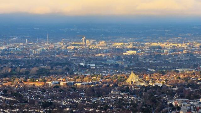 An expansive aerial view of Dublin city at sunset, with warm golden light illuminating the buildings and a soft, hazy sky above. The cityscape stretches into the distance, with the prominent Spire of Dublin visible amongst the various buildings and residential areas.