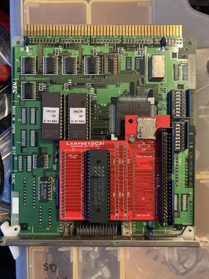 A red board carrying a SCSI controller IC, SD card slot, and internal SCSI header is sandwiched onto the top of a PC-98 SCSI card.