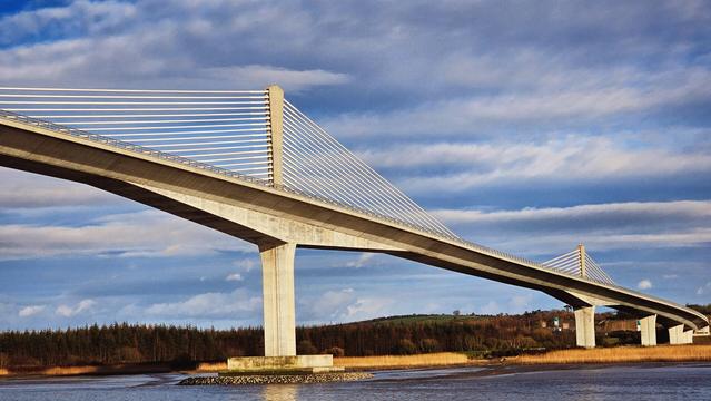 A wide-angle shot of the modern Rose Fitzgerald Kennedy Bridge, a large cable-stayed bridge, spanning a body of water under a dramatic blue sky with white and grey clouds. The bridge's prominent concrete piers and white support cables dominate the frame, connecting the foreground riverbanks to a distant treeline and a small building on the right bank. Reeds and a muddy shoreline are visible along the water's edge in the foreground. The lighting suggests either early morning or late afternoon, casting a warm glow on parts of the bridge structure.