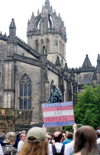 A colour photograph of the Pride march in Edinburgh passing St Gile's Cathedral, which dominates the picture. Benath it is a large crowd of people on the march.

in the centre of the image is a statue of the economist Adam Smith, below which someone is holding a placard which reads "TRANS RIGHTS ARE HUMAN RIGHTS" in black ink on top of the blue, pink, white, pink and blue stripes of the Transgender Pride flag.

There are also Pride flags hanging from the open tower of the cathdral.