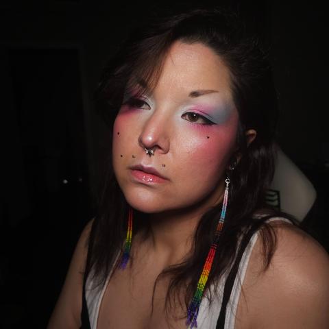 Ashley wearing a pair of long progress pride beaded dangle earrings and eyeshadow in the trans pride colors, from white at the inner corner of the eye, to pink in the middle of the lid, and blue at the outer corner.