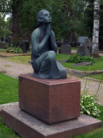 A bronze statue of a kneeling woman with hands pressed together in prayer, sits atop a reddish-brown weathered pedestal in a green summer-time cemetery.