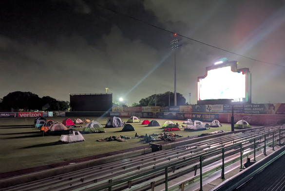 Camping tents pitched on the outfield of professional baseball field  underneath the glow from the scoreboard.