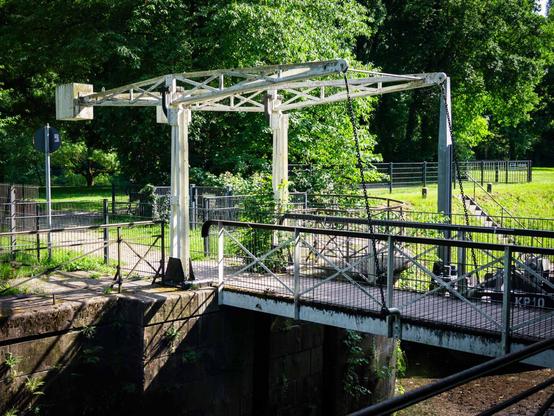 Color photo of a small white metal drawbridge with chains, spanning a narrow water channel in a lush, green park setting. The bridge is surrounded by iron fencing and dappled sunlight.
