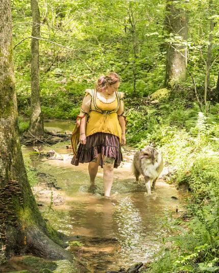 A creek running through a forest, and in the center of it a human and their dog walk towards the camera while smiling at each other, water splashing off their feet as they move through the creek. Sunlight dapples everything. The human is wearing a yellow flowing shirt and red skirt that billows behind as they move, and fairy sleeves cuffed on their arms in yellows and reds. The dog is a fawn & white australian shepherd.