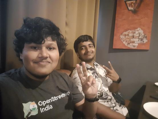 2 men sitting inside a restaurant in Siliguri, facing the camera, raising one hand doing the "V" gesture. The person in the left of the picture is wearing a grey OpenStreetMap India T-shirt.