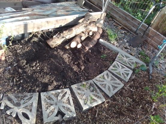 Signs of digging in the bed between the greenhouse extension and the curved path of laid screen blocks. A curved chunk of log mostly buried following inside that curve, a couple more chunks left laying.